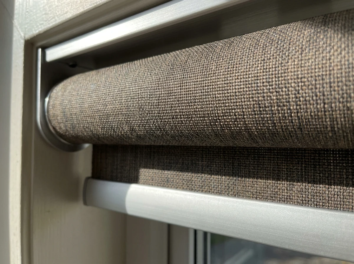 Close-up of a double roller blind in City Bowl — smooth flat woven sunscreen and blockout fabric wound on aluminium tubes with bracket hardware, tobacco and carbon tones